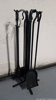 Photo showing the entire set of fireplace tools with the stand. Four tools are hanging from the stand including a poker, tongs, a brush, and a shovel. The set is black metal with a simple design.