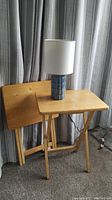 Pair of wooden folding tables with one standing and one folded; ceramic table lamp with blue and white pattern and white cloth shade on standing table