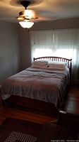 Photo of the full sized wooden framed bed installed in a bedroom, showing the headboard with vertical slats, bedding, and ceiling fan above.