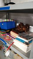 Photo showing blue storage box with striped lid, wooden organizer, stack of books, DVDs including The Family Man, small metal tin, and tape dispenser on lower shelf