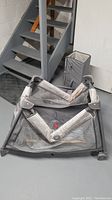 Gray Graco Pack N Play playard unfolded on basement floor, showing mesh side panels and padded frame supports.