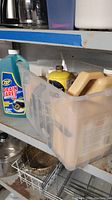 Plastic bin on metal shelving containing multiple household maintenance bottles including Zep Drain Care, yellow rust remover can, caulk gun, and other chemical containers.