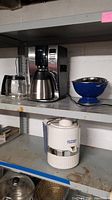 Jack LaLanne Power Juicer, white color, shows cracking and discoloration, chrome band around middle section. Mr. Coffee digital coffee maker with black and silver finish and stainless steel thermal carafe. Blue and silver metal strainers on shelf alongside coffee maker.