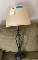 Full view of table lamp showing dark metal base with open geometric design and beige fabric shade, sitting on a green couch arm.