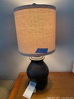 Smaller tabletop lamp with dark bulbous base and beige burlap-style cylindrical lampshade with dark trim.