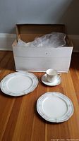 Box with four pieces of Noritake ivory china including two plates, one cup, and one saucer on a wooden floor.