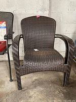 Front view of one dark brown wicker patio chair showing curved armrests and woven seat and backrest against a white textured wall.