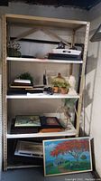 Photo of metal shelving unit with multiple shelves containing picture frames with artwork, glass dome cloche, small pots, stacked books, and other decorative items.