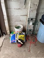 Gardening supplies and equipment including various bags of gardening products, large white bucket, two metal plant stands and a can of red brick paint, all arranged against a cinder block wall in a garage area.