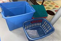 View of large blue plastic storage tote, smaller green tote with lid, blue plastic laundry hamper basket, and two round ceramic pottery planters.