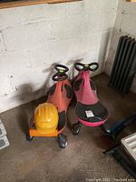 Pair of red and pink kids ride-on scooters next to a yellow kids utility hat and dominoes board game box in a garage corner on a concrete floor.