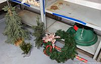 Wide view showing three mini artificial Christmas trees, a green wreath with a decorative bow, two rolls of Christmas wrapping paper, and a green plastic tree stand on a basement floor.