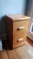 Front view of wooden two-drawer file cabinet against wall and hardwood floor