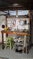 Wooden workbench with pegboard attached to masonry basement wall, worn from use. Various tools seen around but not included in sale. Green wooden stool visible under workbench.