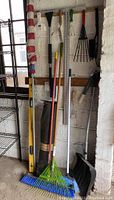 Full view of yard and garden tools hung and standing against a white brick wall. Visible tools include rakes with green and blue tines, a black shovel, a yellow spirit level, and a black rolled mat.
