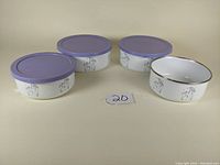 Four enamel over steel food storage bowls with three purple lids attached. One bowl is missing its lid. Bowls feature Corelle Shadow Iris floral pattern with black rim.