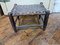 Side view of antique wooden footstool showing woven cane top and rounded legs with crossbar support.