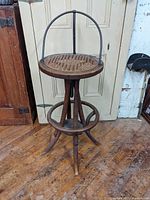 Front view showing the full antique stool with bent wood legs, cane seat, and wrought iron back brace arch