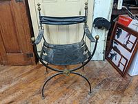 Front angled view of the iron and leather Savonarola chair showing worn black leather seat and brass finials on armrests and center crossing piece.