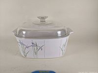 Front angle photo showing the white deep Corning Ware casserole dish with Shadow Iris floral pattern and clear glass lid with knob.