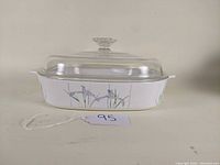 Front side view of a white shallow oval Corning Ware casserole with a clear glass lid. The dish displays the Shadow Iris pattern with purple and green floral design.