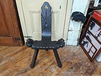 Front view of the black painted carved folk art three-legged chair showing seat, legs, backrest with sunburst carving.