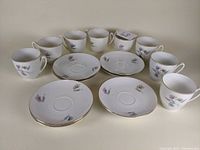 8 white porcelain teacups and matching saucers arranged in rows, floral Cornelia pattern with gold rims.