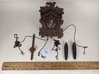 Vintage carved walnut cuckoo clock displayed with its pendulum, two pinecone weights, and chains stretched out beside it, all shown next to a measuring ruler for scale.