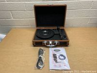 Open suitcase-style record player showing black turntable and soul, control knobs and buttons on the right, USB cable and instruction manual beside it on the table.