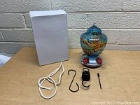 Blown glass hummingbird feeder displayed alongside its original white box and hanging accessories including a rope, metal S-hook, coil spring, and metal rod.