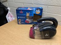Bissell Pet Hair Eraser handheld vacuum cleaner with packaging box behind it on a wooden surface.