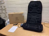 Photo of the Comfier massage seat cushion displayed on a wooden table next to its original box, remote control, and power cable.