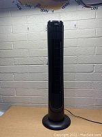 Front view of black DREO tower oscillating fan on tabletop against light gray brick wall background.