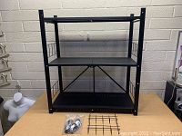 Foldable black metal storage shelving unit with three perforated shelves, mesh wire sides, and included caster wheels and metal grid parts displayed in front.