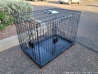 Side view of the black foldable metal wire dog crate with double doors and plastic tray base, set outdoors on pavement near some landscaping.