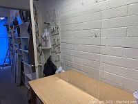 Clear plexiglass sneeze guard standing upright on wooden table against brick wall background. Pass-through slot visible near bottom center.