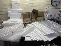 Large assortment of packing materials: white foam sheets, foam corner protectors, two brown cardboard boxes, and bubble wrap laid out on a table.