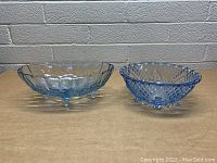 Two vintage blue glass dishes on a wooden surface against a brick wall. Oval dish on left with scalloped edges, round dish on right with patterned design.