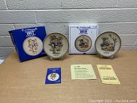 Two vintage M.J. Hummel plates dated 1977 and 1979 displayed on stands with original boxes and authenticity certificates shown on table against a grey brick wall background.