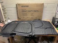 Set of four black heavy duty deep dish floor mats laid out on a table with the original box standing behind. Mats have textured non-slip surfaces and raised edges to trap dirt and liquids. Each mat is labeled 'Motor Trend'.