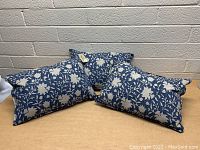 Three rectangular blue and white floral patterned throw pillows displayed on a wooden surface against a brick wall background, all new condition.