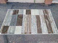 Top-down view of rectangular small area rug laid flat on asphalt, showing full pattern of abstract blocks in neutral tones.