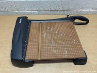 X-ACTO paper cutter showing top view with wooden base, clear marked overlay, black cutting arm and handle, and ruler guard.