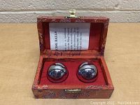 Two reflective metallic Baoding Chiming Balls seated in a red silk-lined box with an open lid showing a leaflet inside.