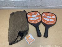 Photo showing the two pickleball paddles, black mesh carry bag, and square informational card on a table.