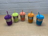 Front view of five stainless steel tumblers featuring silicone sleeves and matching lids and straws in purple, light green, peach, orange, and blue colors against a neutral background.
