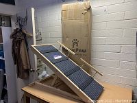 Photo showing the adjustable wooden pet ramp with blue-gray carpet and movable side rails, with original RamPaw packaging in background.