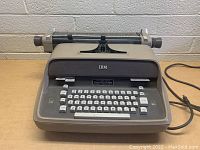 Front view of the vintage IBM electric typewriter, showing keyboard, platen roller, and IBM logo.