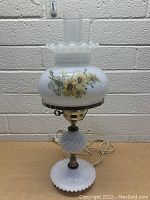 Full front view of the vintage milk glass table lamp showing floral decorated milk glass shade with clear glass chimney, beaded edge base, and ornate twisted milk glass stem section. The lamp is plugged in and switched off.