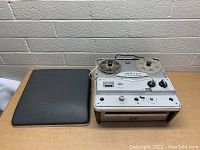 Front diagonal view of the Pelouze Magnetics reel to reel tape recorder with one reel and a cover placed to the left on a wooden surface against a brick wall.
